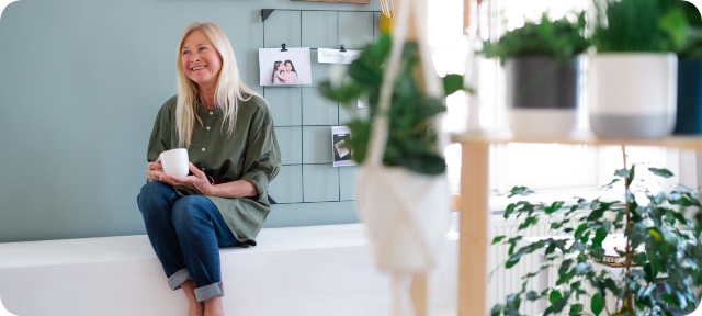 Woman sits in living room, holds coffee mug and smiles.